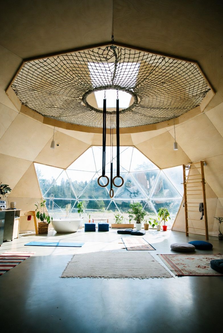 The one-room dome, whole: a rope net loft under a central oculus, gymnastic rings, a freestanding tub, plants along the glazed band, and wall bars — all in a single daylit room.